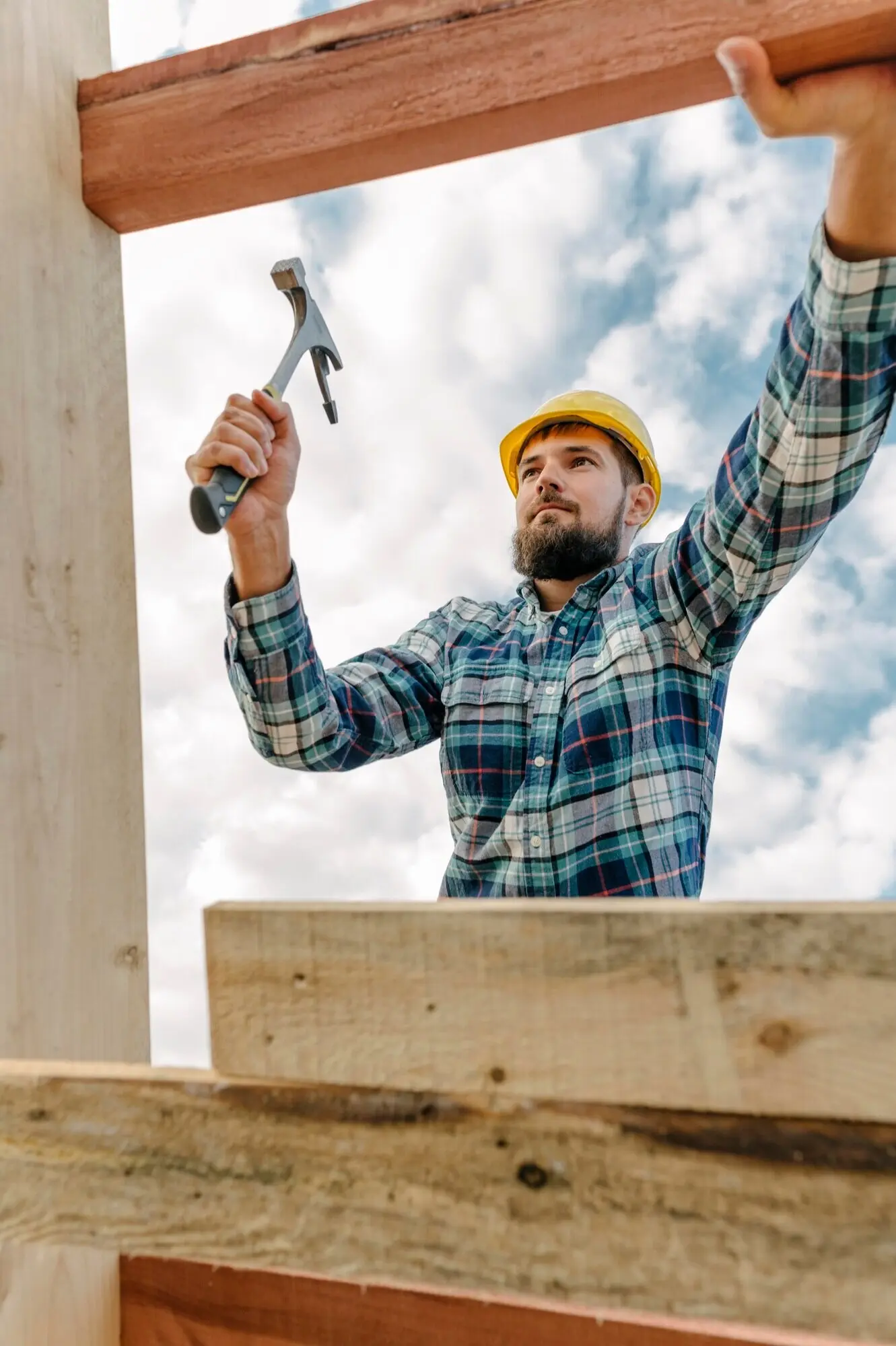 A construction worker wearing a hard hat and holding a hammer is building the roof of the house.
